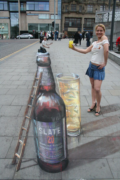 3D sidewalk illusion of a giant Slate bottle and a glass, with the artist posing on the top and looking tiny