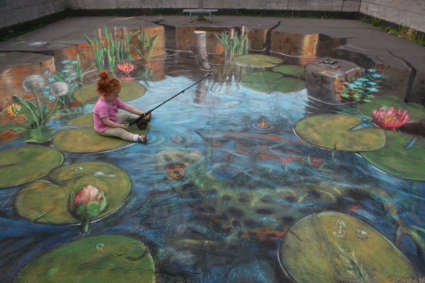 3D chalk illusion of a pond with water lillies and a newt underwater, with a real little girl trying to fish it