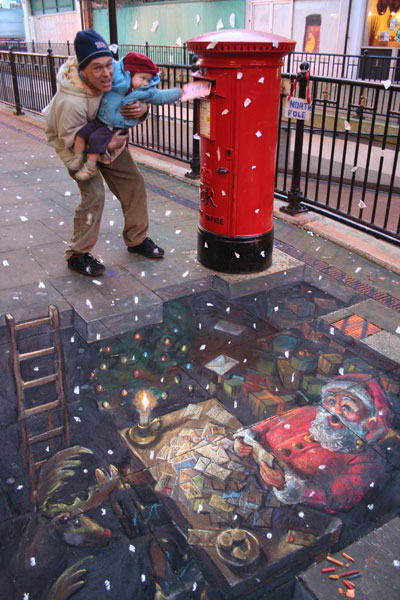 3D chalk art of Santa's underground workshop while above ground a real toddler puts a letter in a real letterbox , supposedly falling straight into the workshop