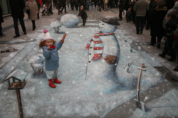 3D pavement drawing of a snowman with a real little girl about to put a carrott on its face